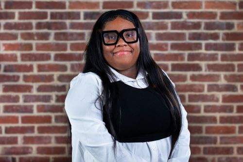 Nalisa Jacobs, a young Black woman, stands in front of a brick background. She is wearing thick rimmed glasses, a white blouse, and a sweater vest.